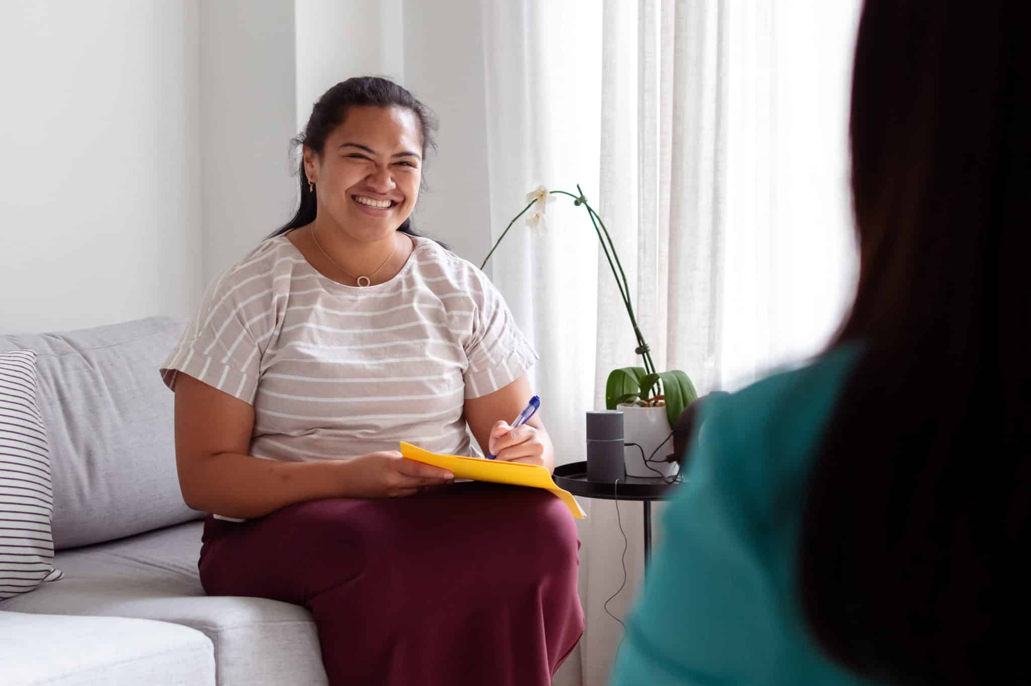 A positive young therapist of Pacific Islander descent smiles while talking with an unidentifiable female client during a psychotherapy session.